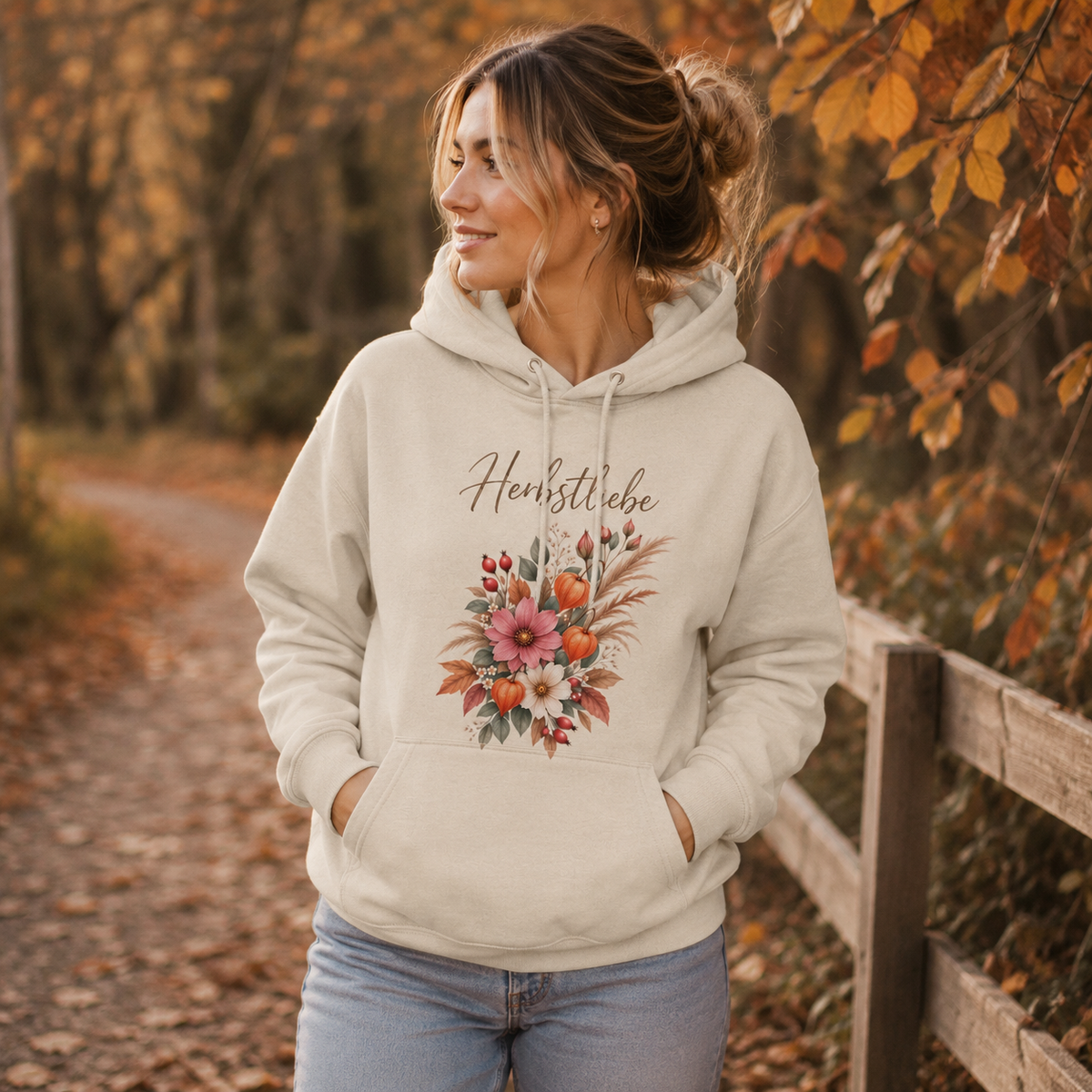 Frau trägt cremefarbenen Nomavé Unisex Hoodie Herbstliebe mit floralem Herbstmotiv auf einem herbstlichen Waldweg