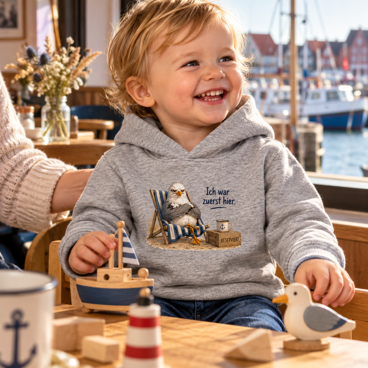 Kleinkind in graumeliertem Baby-Hoodie mit Frontmotiv Möwe Alpha in einem Hafencafé.