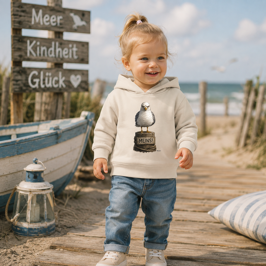Kleinkind trägt den Baby Organic Hoodie „Möwe mit Meinung – MEINS!“ von Nomavé auf einem Holzsteg am Strand. Der cremefarbene Hoodie mit der grimmigen Möwe auf Poller sorgt für einen maritimen Küstenlook mit norddeutschem Humor.