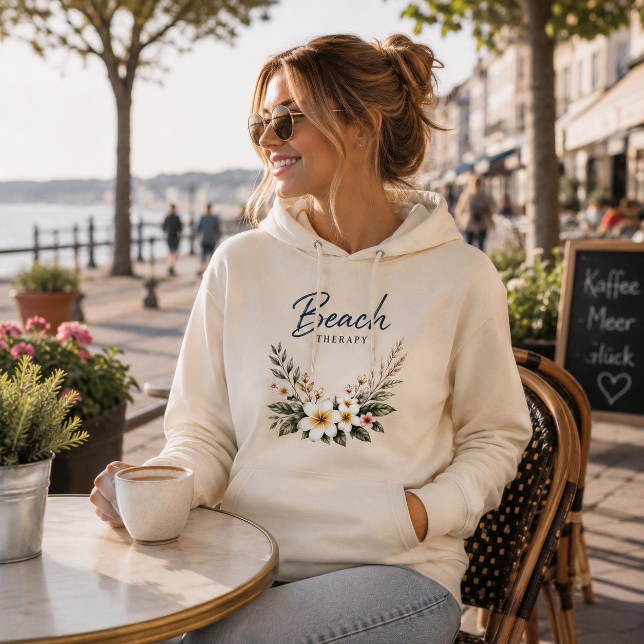 Junge Frau trägt einen cremefarbenen Hoodie mit dem floralen Motiv „Beach Therapy“ von Nomavé und sitzt entspannt in einem Café an einer maritimen Promenade. Das hochwertige Blumen-Design vermittelt sommerliche Leichtigkeit, Küstengefühl und entspannte Beach-Vibes.