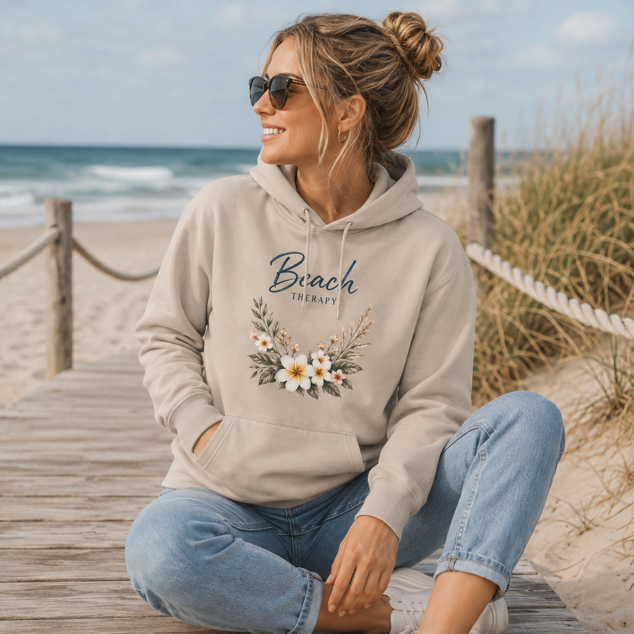 Frau im beigen Beach Therapy Hoodie mit floralem Motiv auf einem Holzsteg am Strand