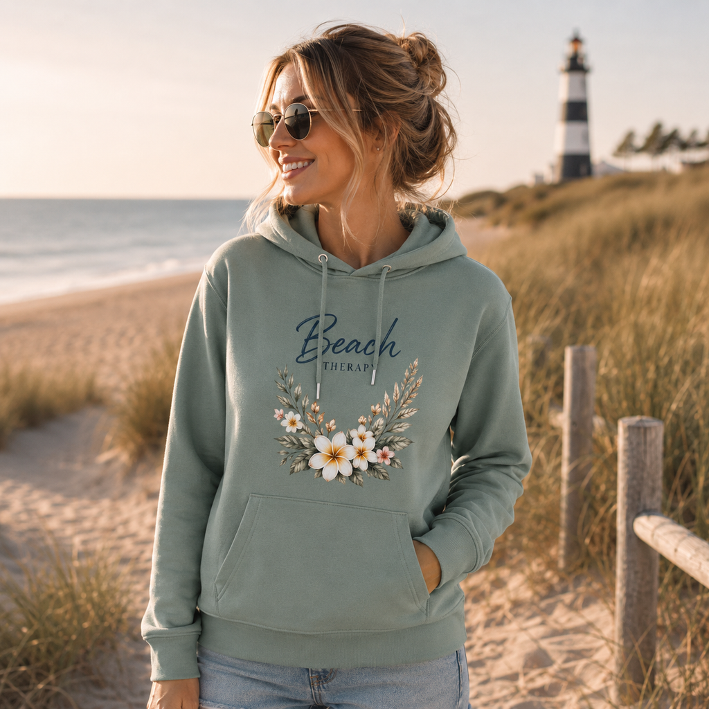 Junge Frau trägt einen salbeigrünen Hoodie mit dem floralen Motiv „Beach Therapy“ von Nomavé. Das hochwertige Blumen-Design wird in einer warmen Strandkulisse mit Dünen, Meer und Leuchtturm präsentiert und vermittelt sommerliches Küstengefühl und entspannte Beach-Vibes.