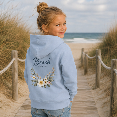 Kind im hellblauen Beach Therapy Hoodie mit floralem Rückendruck auf einem Holzsteg am Strand