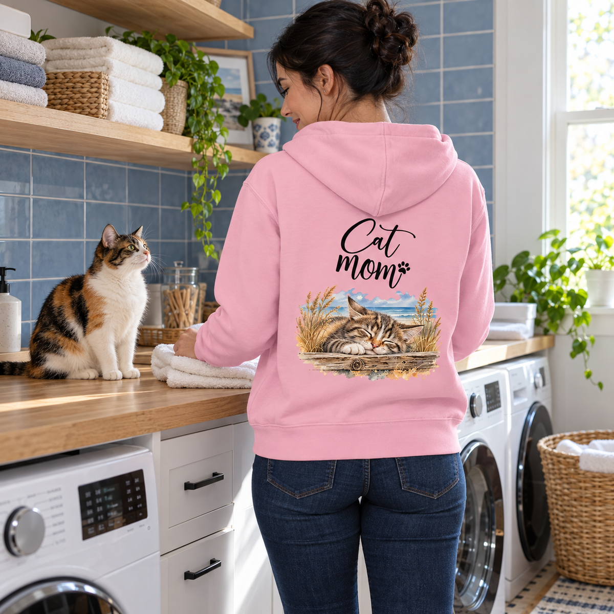 Cat Mom Bio-Hoodie in Cotton Pink mit Rückenprint, Frau faltet Handtücher neben einer dreifarbigen Katze im Hauswirtschaftsraum.