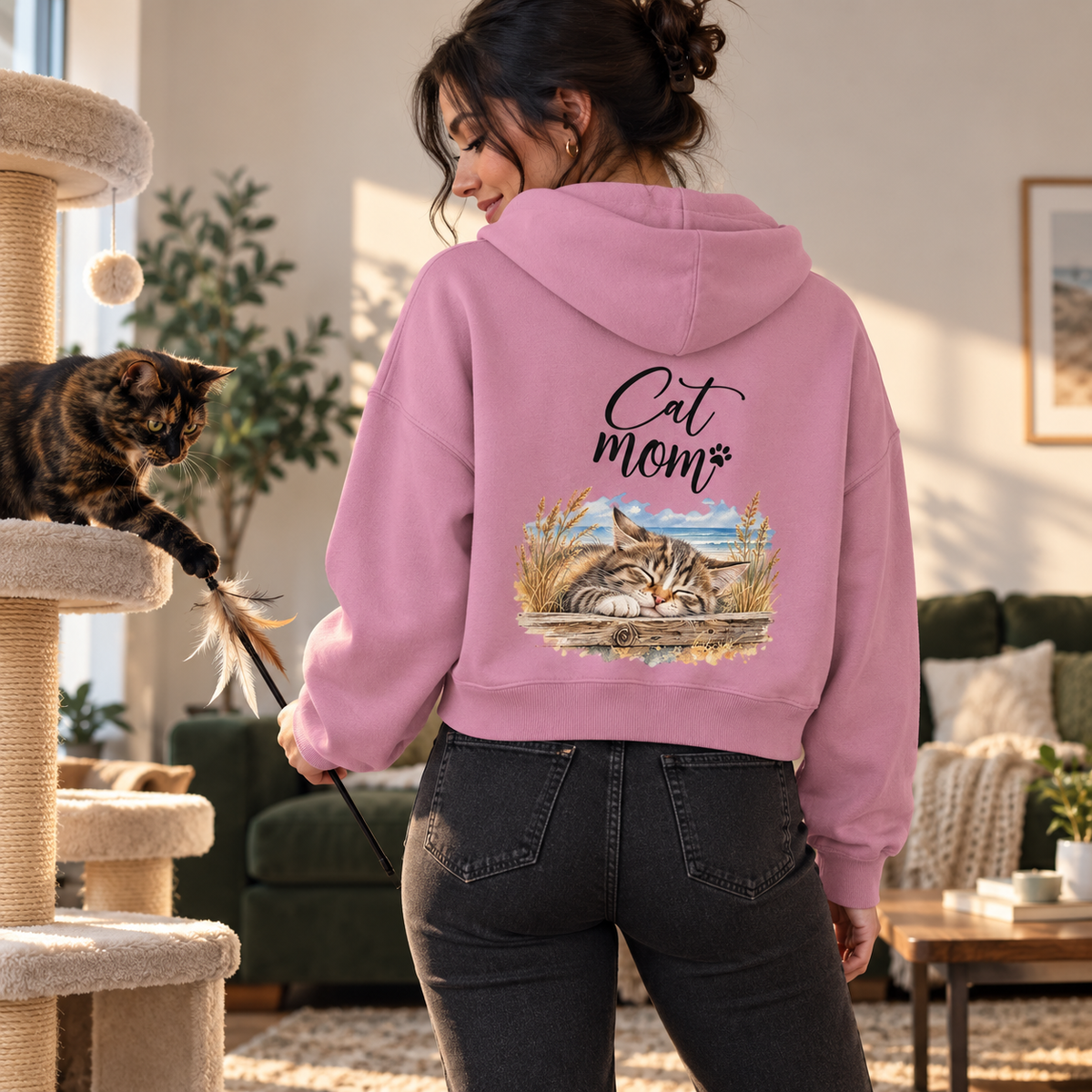 Cat Mom Damen Bio-Boxcut-Hoodie in Bubble Pink mit Rückenprint, Frau spielt am Kratzbaum mit einer Schildpattkatze.
