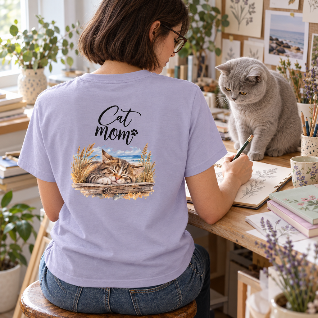 Cat Mom Damen Bio-T-Shirt in Lavender mit Rückenmotiv, Frau zeichnet am Schreibtisch neben einer grauen British-Shorthair-Katze.