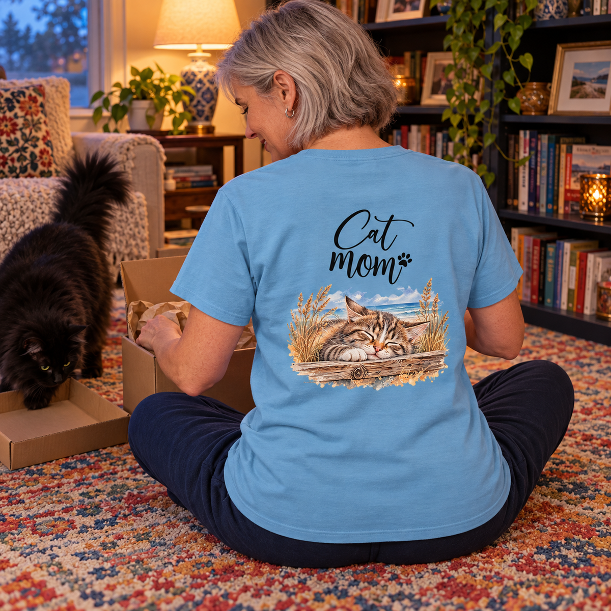 Cat Mom Heavy-Cotton-T-Shirt in Carolina Blue mit Rückenprint, Frau packt am Wohnzimmerboden einen Karton aus, schwarze Langhaarkatze daneben.