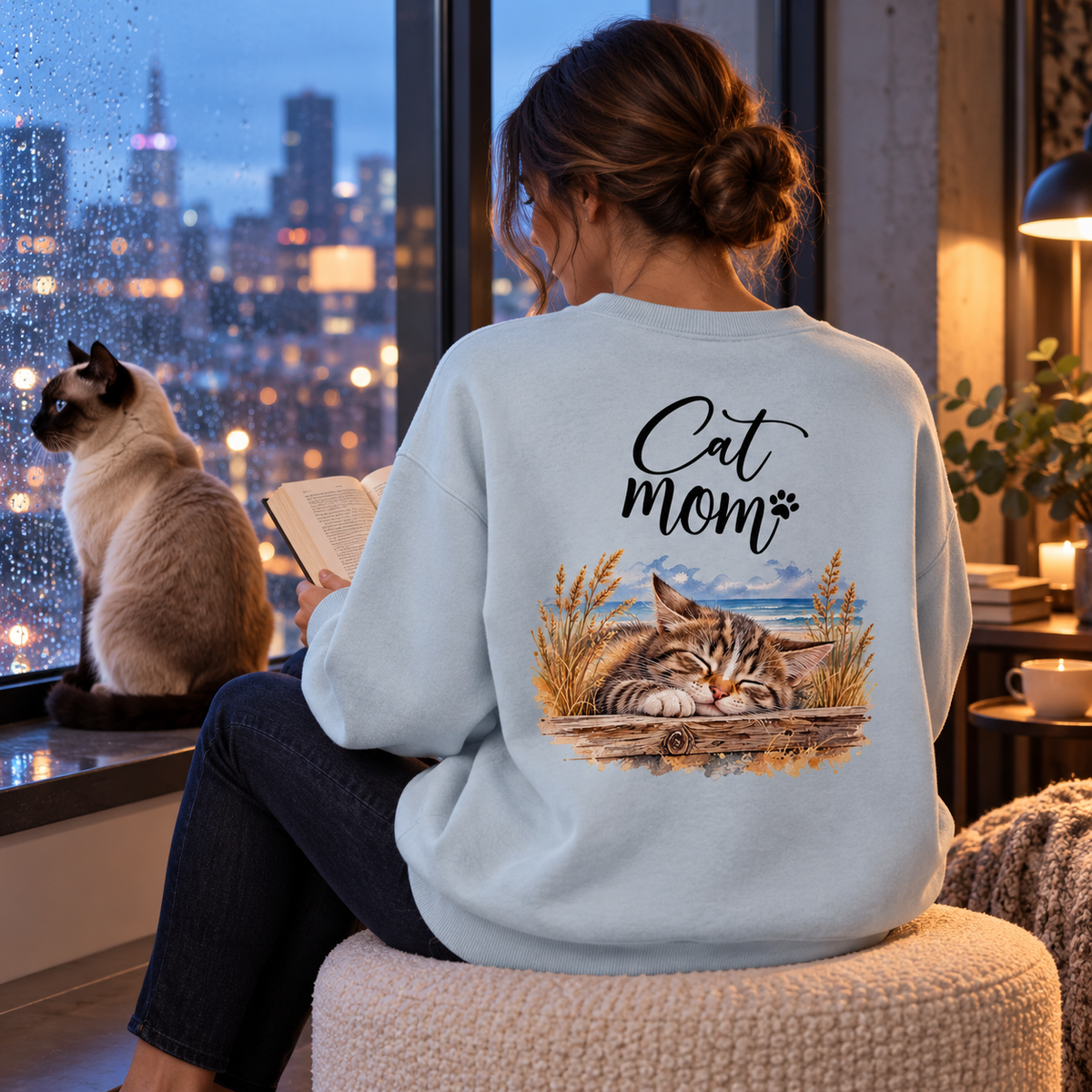 Cat Mom Bio-Oversize-Sweatshirt in Blue Ice mit Rückenprint, Frau liest am regennassen Fenster neben einer Siamkatze im Abendlicht.