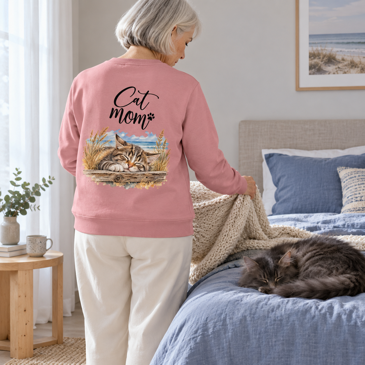 Cat Mom Unisex Bio-Sweatshirt in Canyon Pink mit Rückenprint, Frau faltet eine Decke im Schlafzimmer neben einer schlafenden grauen Langhaarkatze.