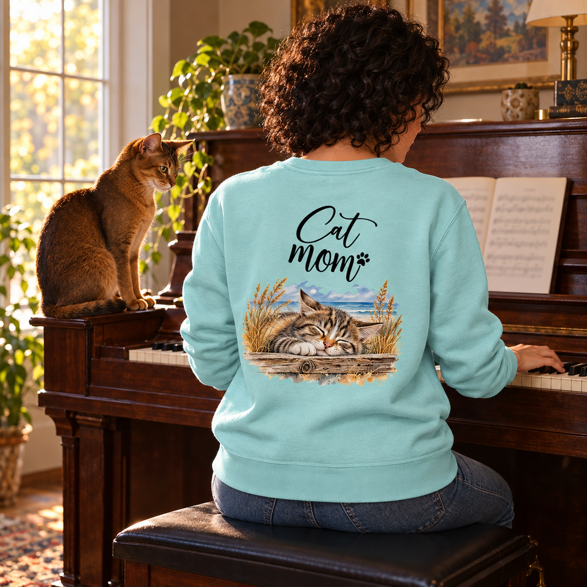 Cat Mom Unisex Bio-Sweatshirt in Caribbean Blue mit Rückenprint, Frau spielt Klavier neben einer Abessinierkatze.