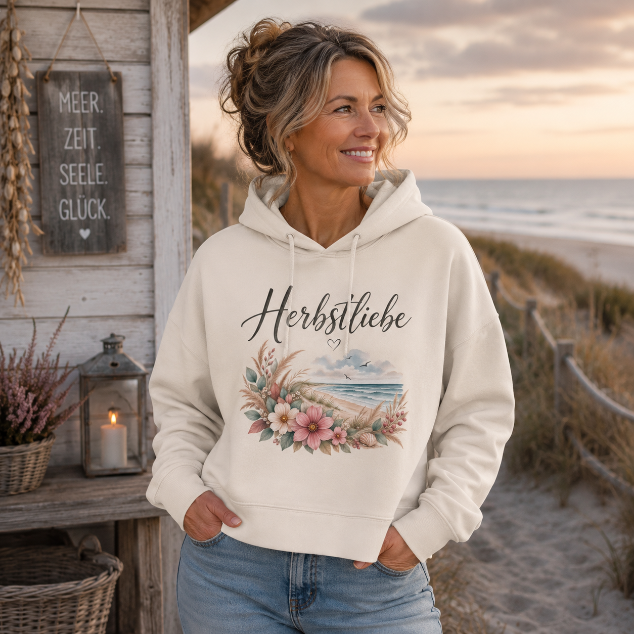 Frau mittleren Alters im cremefarbenen Herbstliebe Boxcut Hoodie am Meer – florales Küstenmotiv der Autumn Coast Kollektion