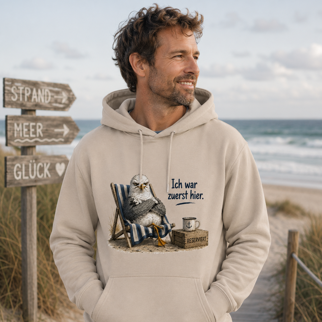 Mann am Strand im naturfarbenen Hoodie mit Möwe und Spruch „Ich war zuerst hier“ aus der Nomavé Küstenhumor Kollektion