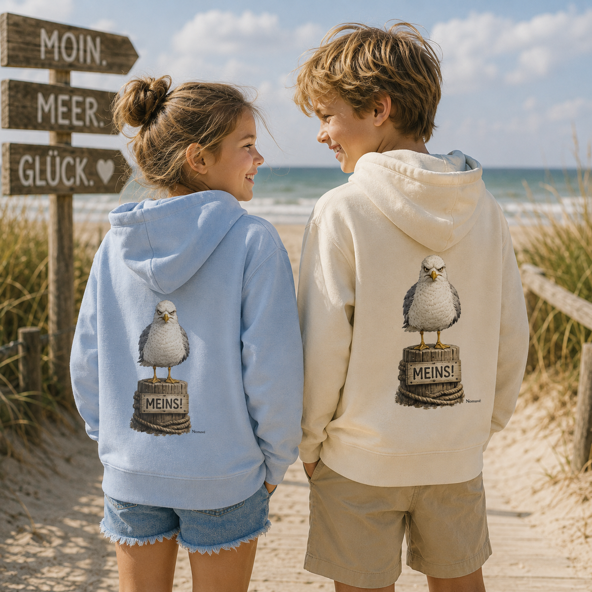 Zwei Kinder tragen Bio-Kinderhoodies von Nomavé in Natur und Hellblau mit dem Motiv „Möwe mit Meinung – MEINS!“ auf dem Rücken und stehen gemeinsam auf einem Strandweg an der Ostsee.