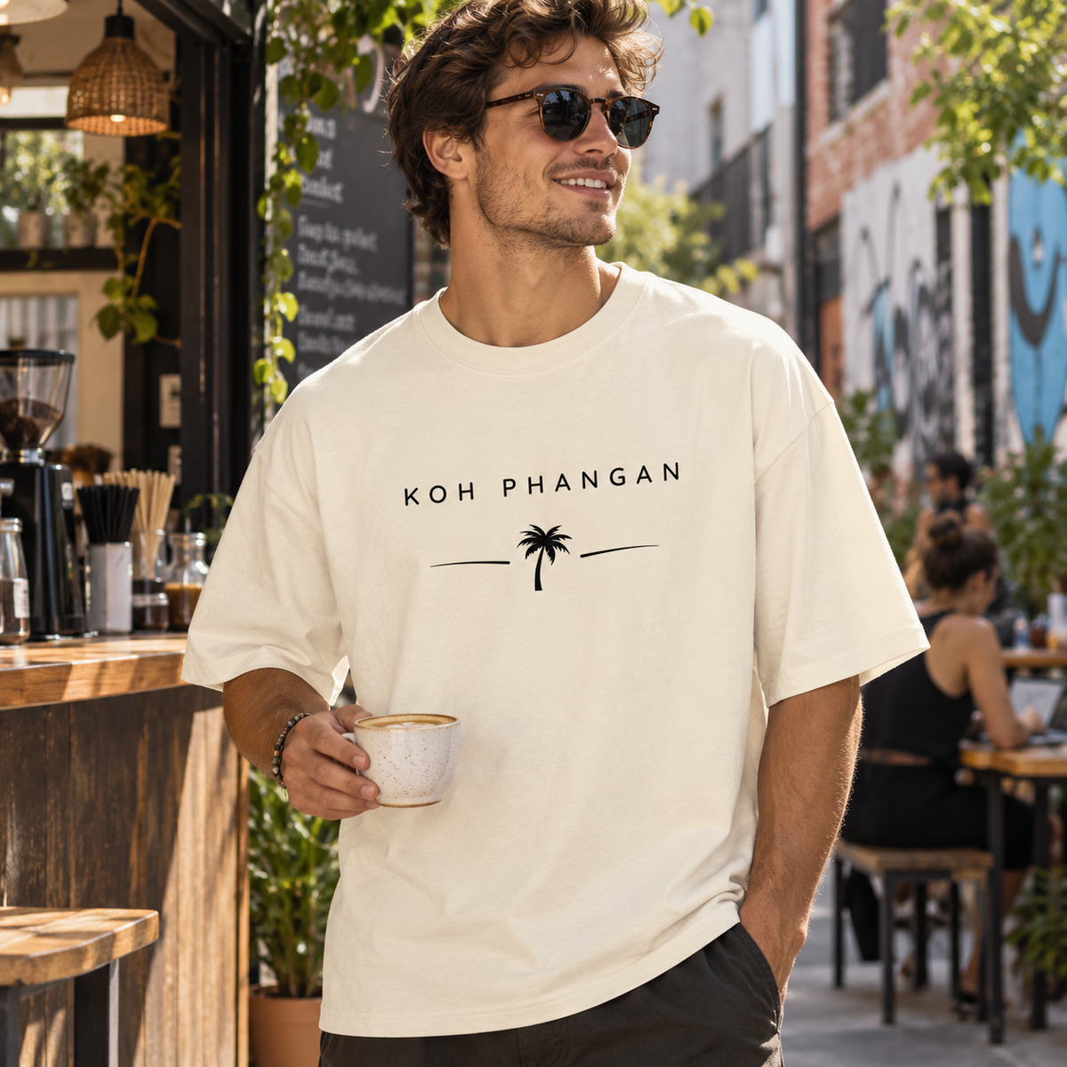 Mann im Bio Oversize T-Shirt mit Koh Phangan Minimal Palm Frontprint an einem Stadtcafé