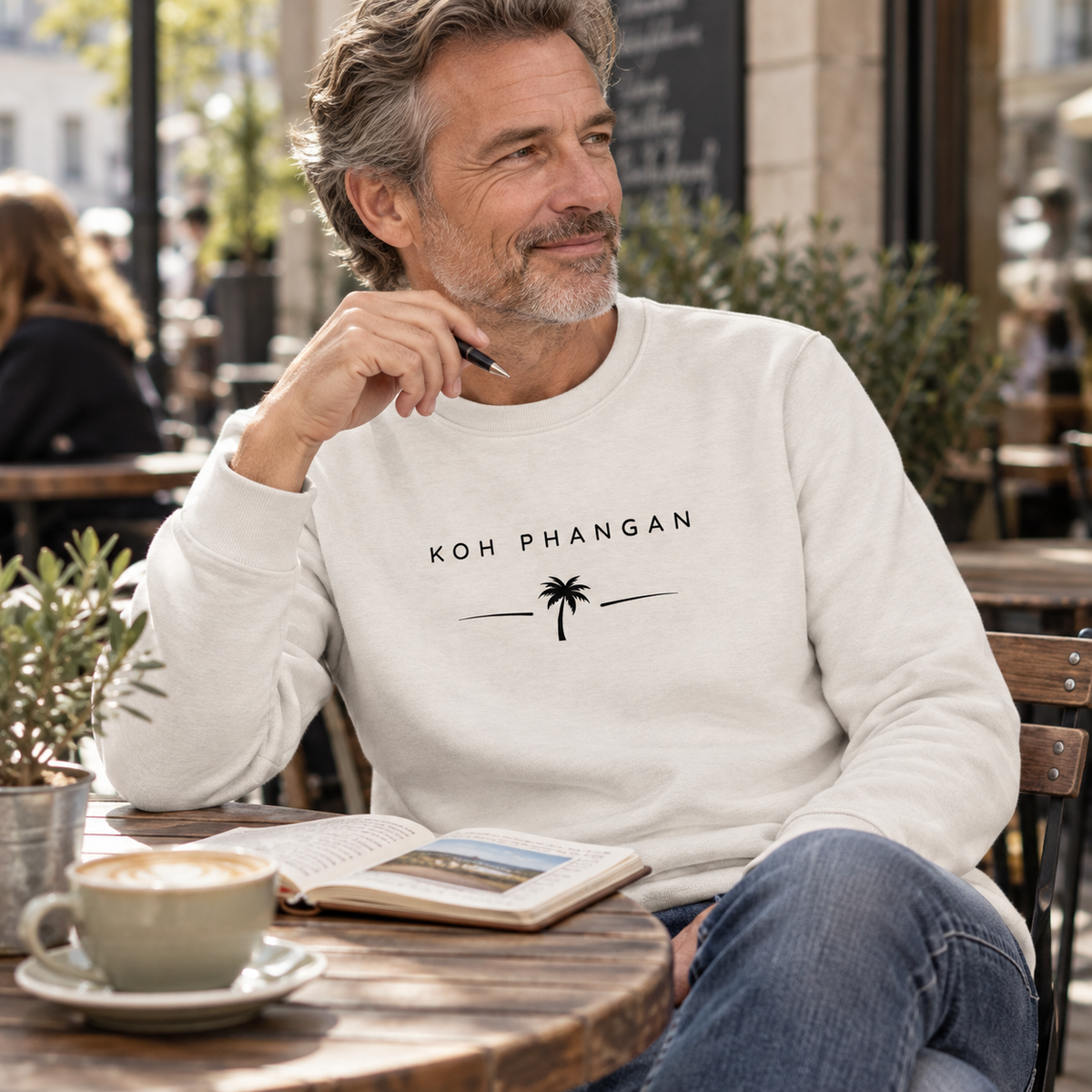 Mann im Bio Unisex Sweatshirt mit Koh Phangan Minimal Palm Motiv an einem Straßencafé