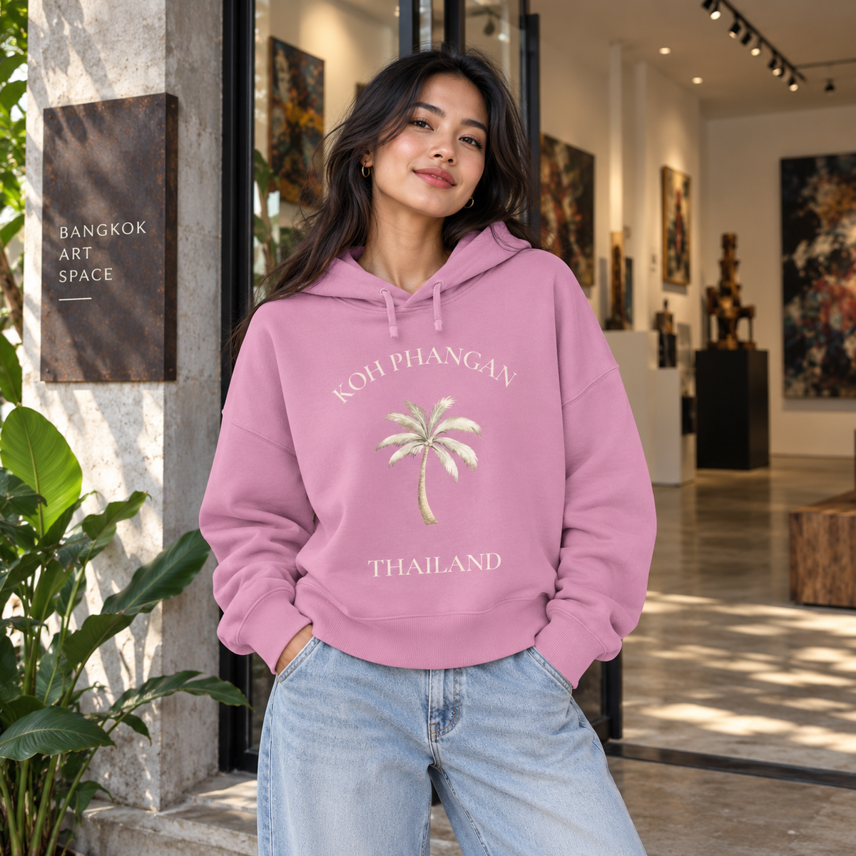 Frau im Bubble Pink Damen Bio Boxcut Hoodie mit Koh Phangan Mini Palm Frontprint vor einer Galerie in Bangkok