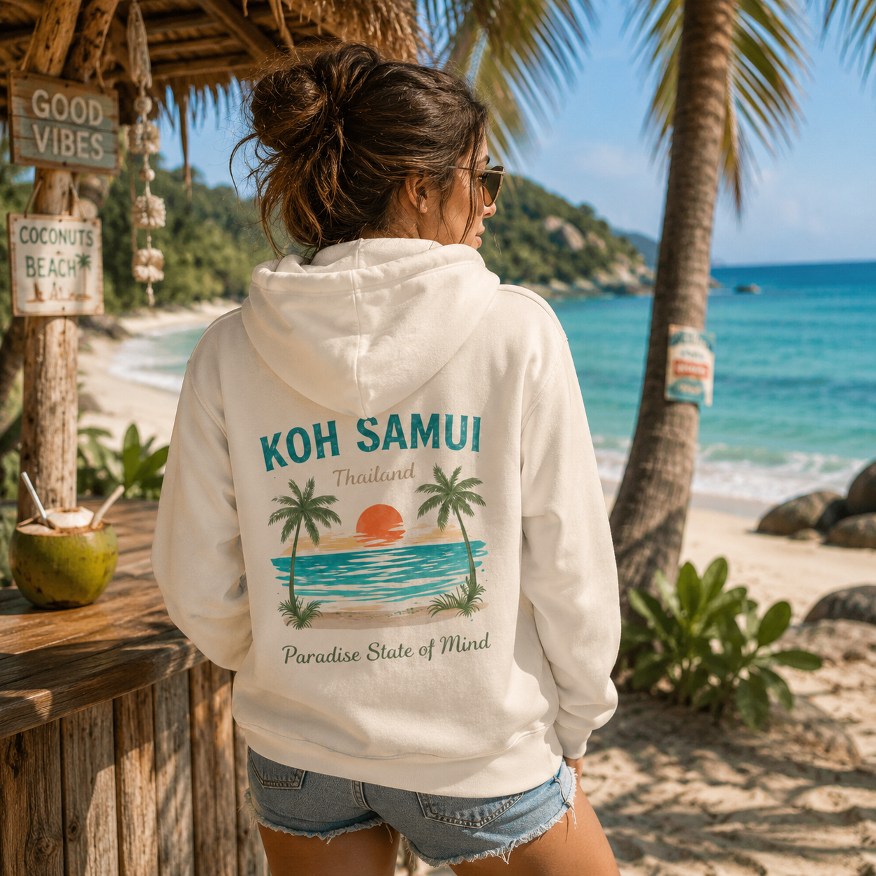 Frau trägt die Bio Kapuzenjacke mit Reißverschluss „Koh Samui – Paradise State of Mind“ mit großem Rückenprint an einer tropischen Beachbar auf Koh Samui in Thailand. Die Vorderseite der Kapuzenjacke ist unbedruckt, das Motiv befindet sich ausschließlich auf der Rückseite.