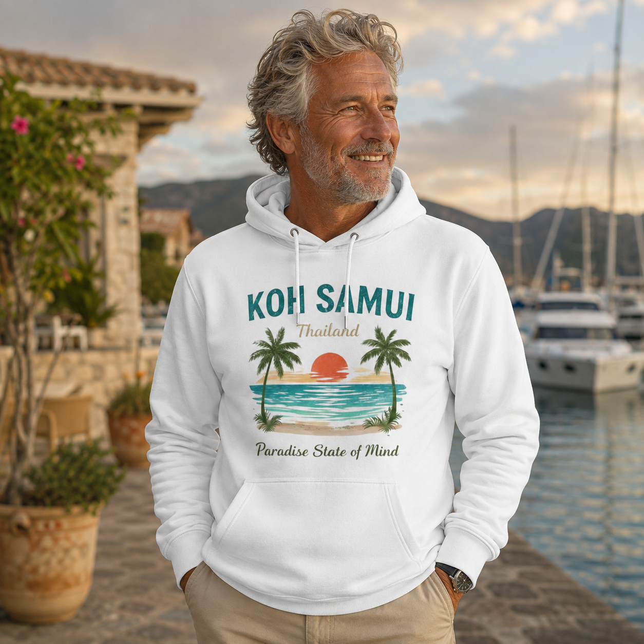 Älterer Mann trägt einen naturweißen Bio Hoodie mit Koh Samui Thailand Motiv an einer tropischen Marina mit Yachten und Abendstimmung.