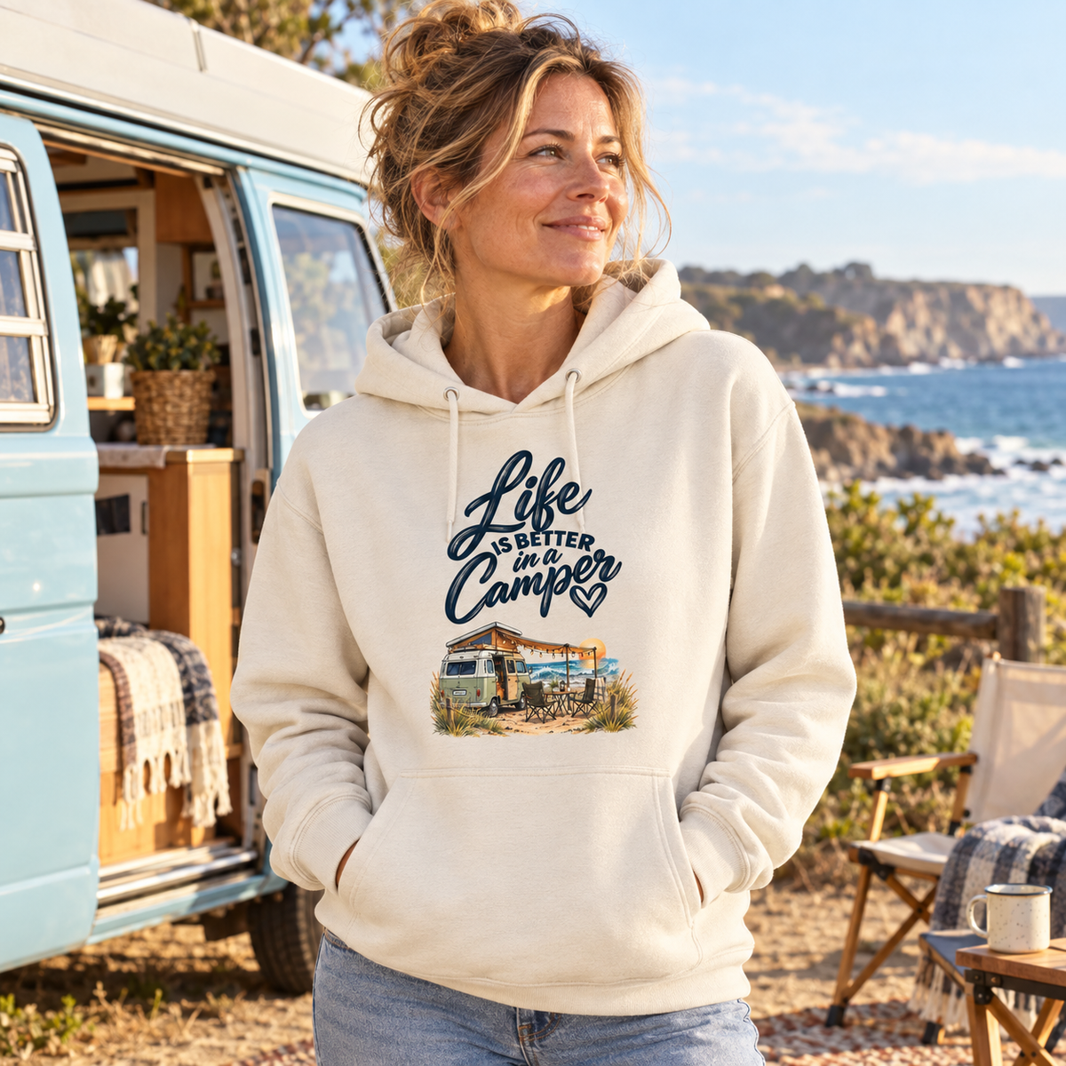 Frau im Bio-Hoodie mit Camper-Motiv vor hellblauem Vintage-Camper an der Küste