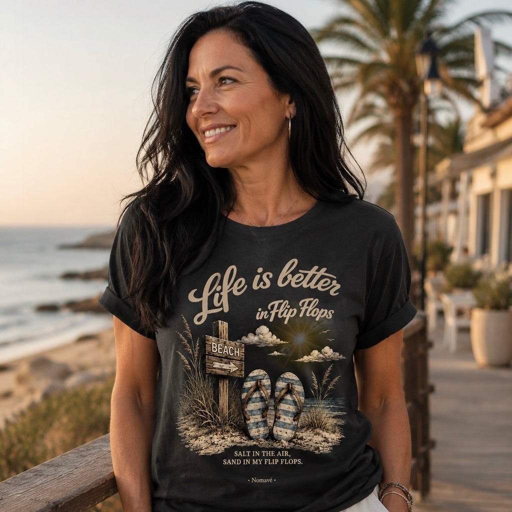 Frau mit langen schwarzen Haaren trägt das dunkelgraue Bio T-Shirt „Life is better in Flip Flops – Dark Edition“ von Nomavé auf einer sonnigen Strandpromenade mit Meer im Hintergrund.