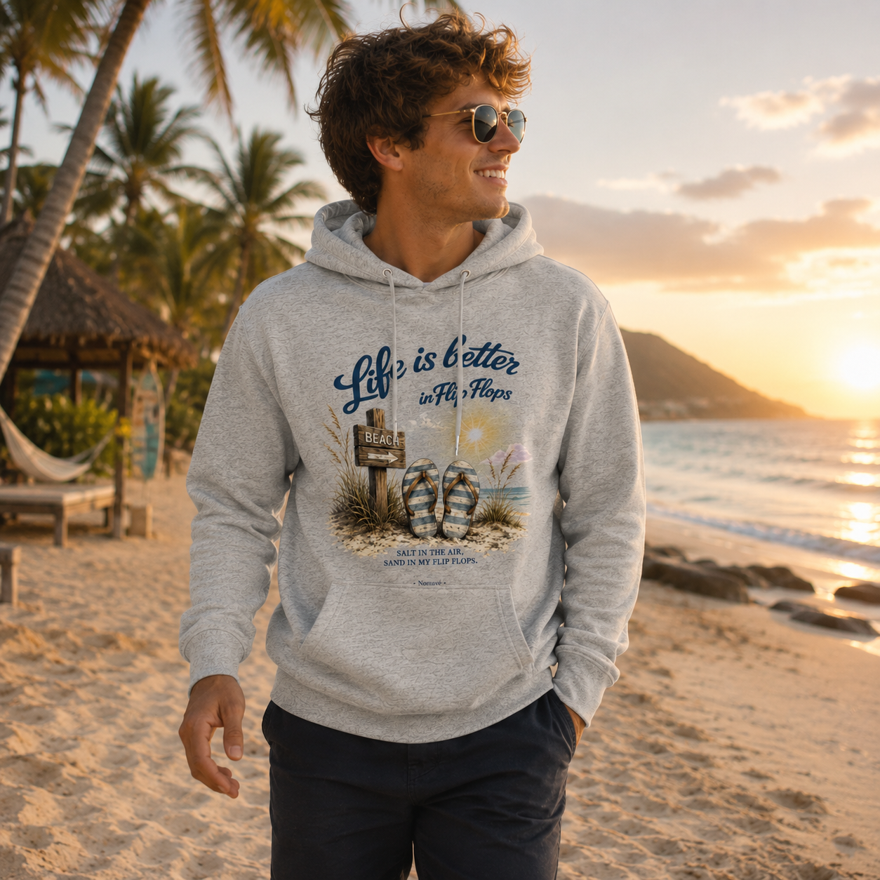 Junger Mann trägt grauen Hoodie „Life is Better in Flip Flops“ und spaziert an einem tropischen Palmenstrand bei Sonnenuntergang.