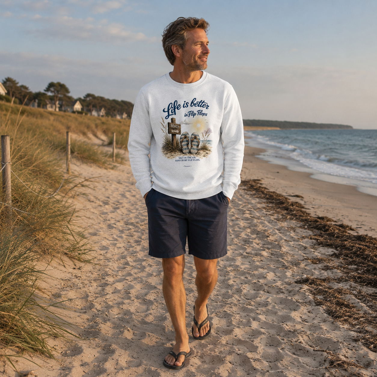Mann trägt weißes Sweatshirt „Life is Better in Flip Flops“ und spaziert in Flip-Flops am Ostseestrand entlang der Dünen.