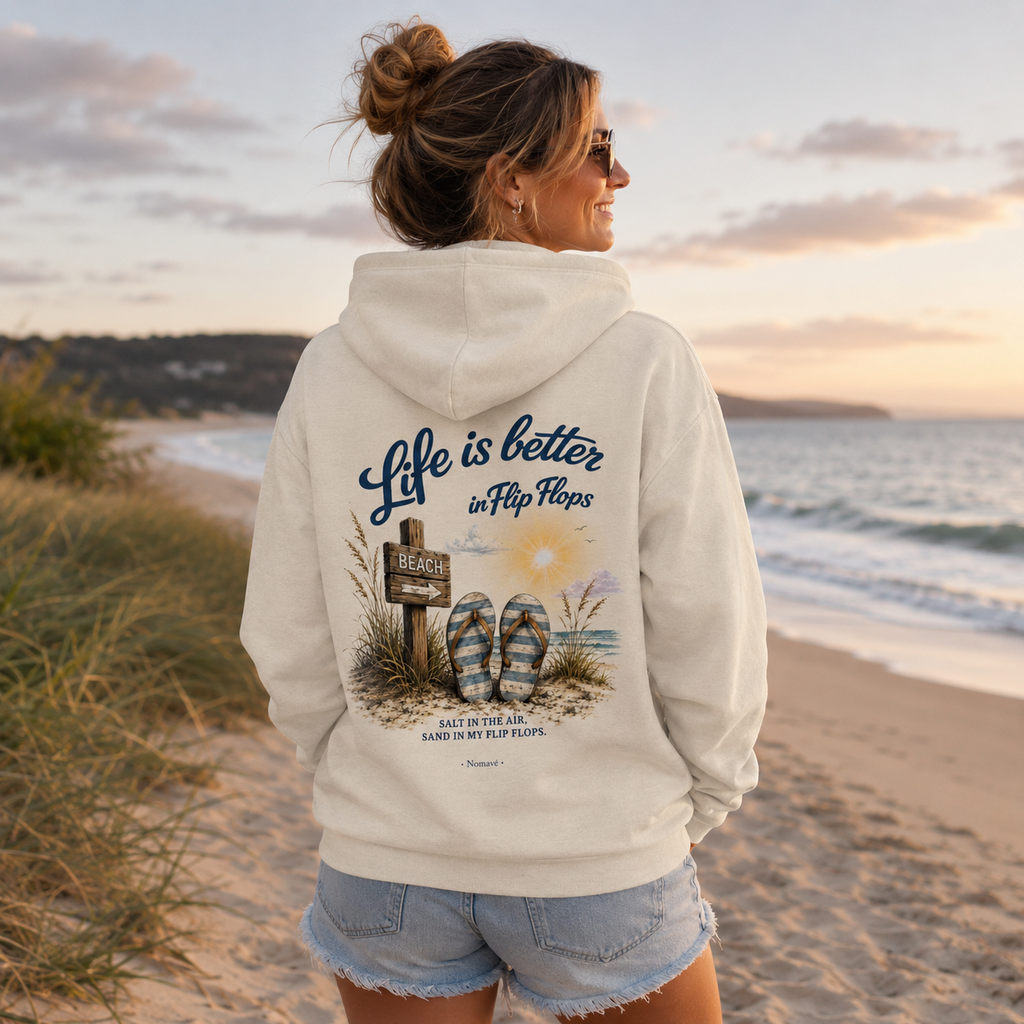 Frau trägt Nomavé Kapuzenjacke mit Reißverschluss und großem Rückenmotiv „Life is Better in Flip Flops“ am Ostseestrand bei Sonnenaufgang.