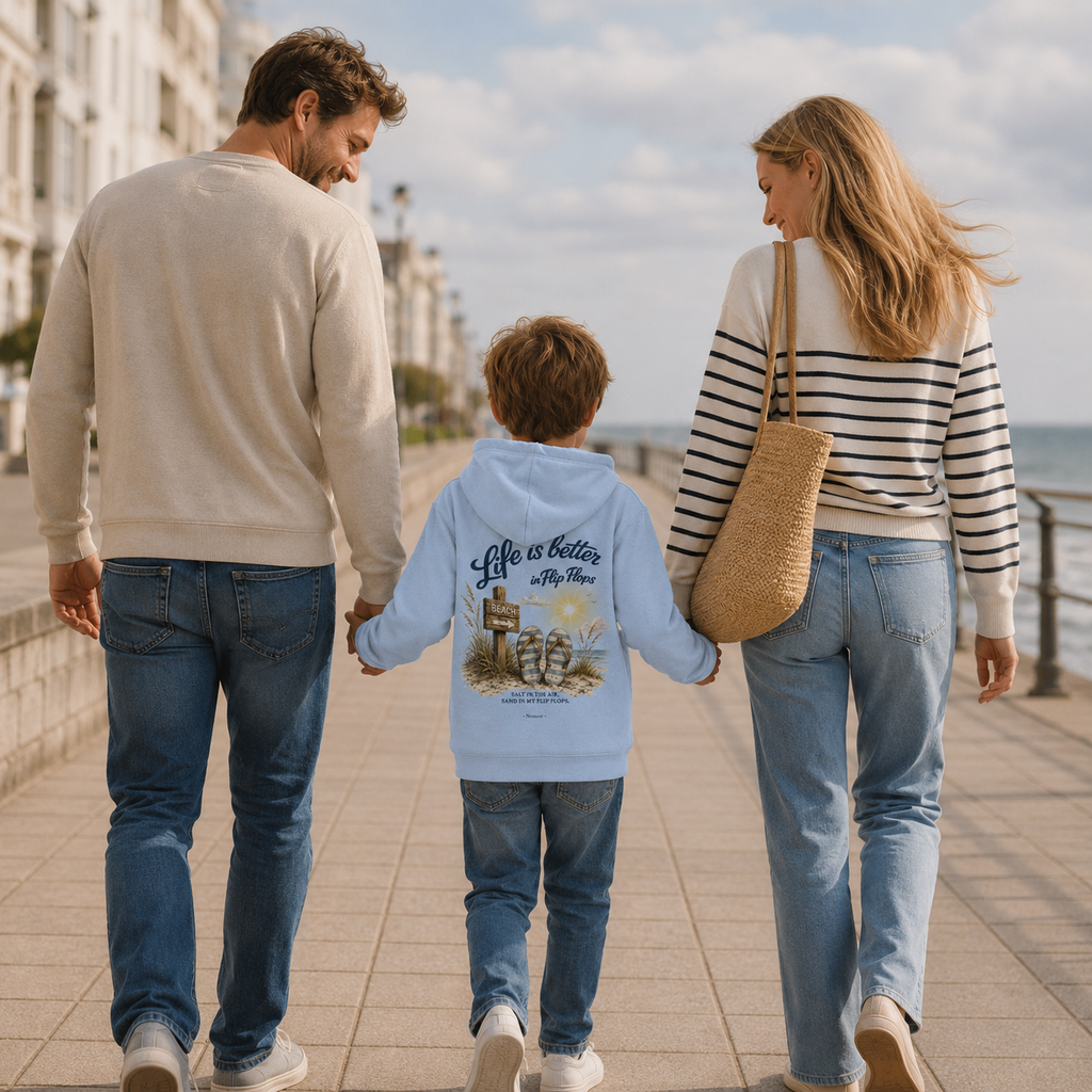 Familie spaziert gemeinsam über die Ostsee-Promenade. Das Kind trägt den Nomavé Kinder-Hoodie „Life is Better in Flip Flops“ mit Rückendruck in Hellblau.