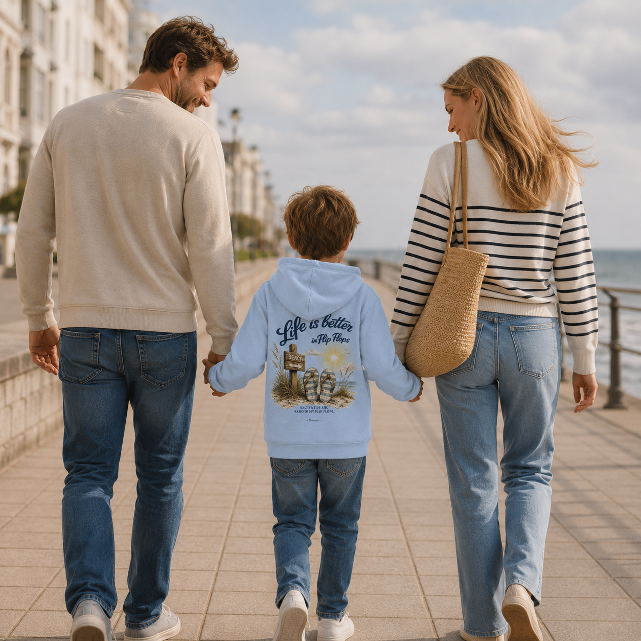 Familie spaziert gemeinsam über die Ostsee-Promenade. Das Kind trägt den Nomavé Kinder-Hoodie „Life is Better in Flip Flops“ mit Rückendruck in Hellblau.