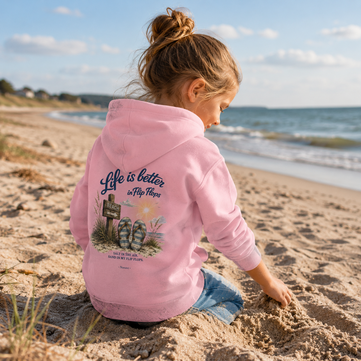 Mädchen spielt im Sand am Ostseestrand und trägt den rosafarbenen Nomavé Kinder-Hoodie „Life is Better in Flip Flops“ mit großem Rückendruck.