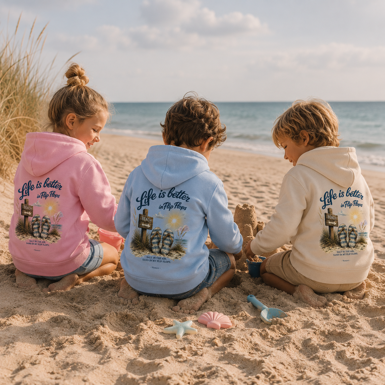 Drei Kinder spielen am Ostseestrand im Sand und tragen Nomavé Kinder-Hoodies „Life is Better in Flip Flops“ mit Rückendruck in Rosa, Hellblau und Naturweiß.