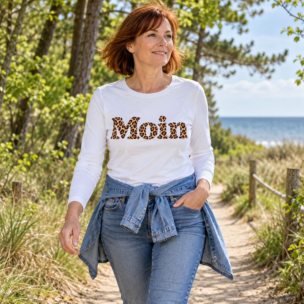 Frau im weißen Bio Langarmshirt mit lesbarem Moin-Leoprint auf einem Küstenparkweg
