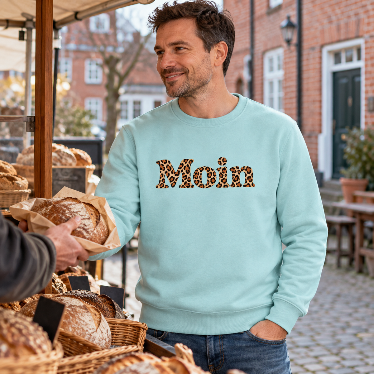 Mann im Bio Unisex Sweatshirt in Caribbean Blue mit Moin-Karamell-Leoprint als Frontprint am Bäckereistand