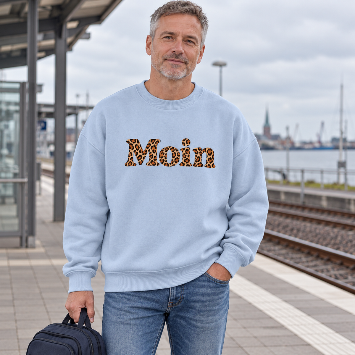 Mann im Bio Oversize Sweatshirt in Blue Ice mit Moin-Karamell-Leoprint als Frontprint am Bahnhof