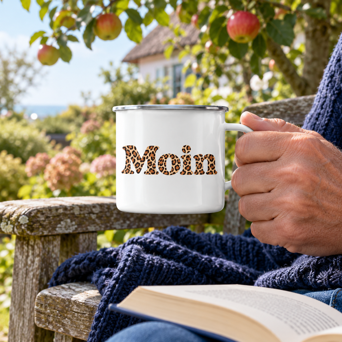 Hand hält die weiße Moin-Leo-Emaille-Tasse mit Silberrand und Karamell-Leoprint an einer Gartenbank
