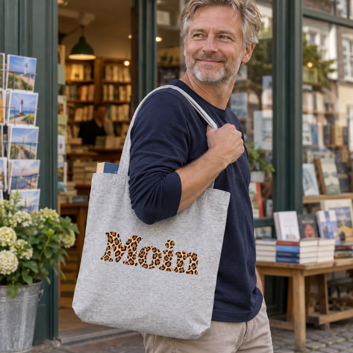 Mann trägt die Tote-Bag in Heather Grey mit Moin-Karamell-Leoprint an langen Henkeln vor einem Buchladen