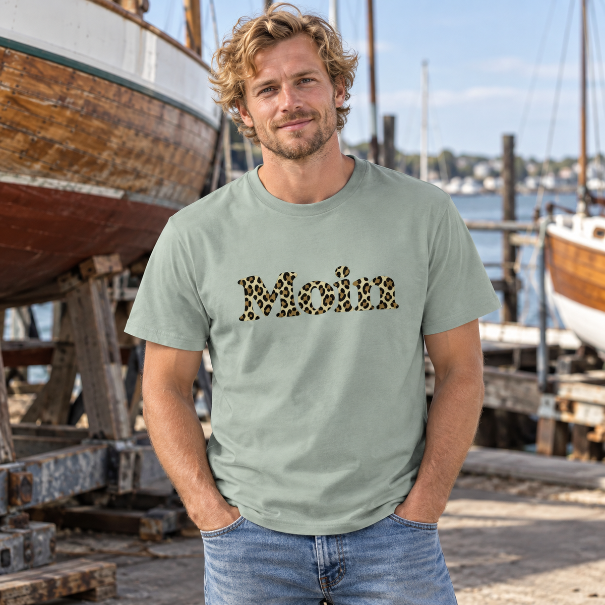 Mann im Bio Unisex T-Shirt in Aloe mit Moin-Leoprint der Sage-Green-Edition als Frontprint an einer Bootswerft