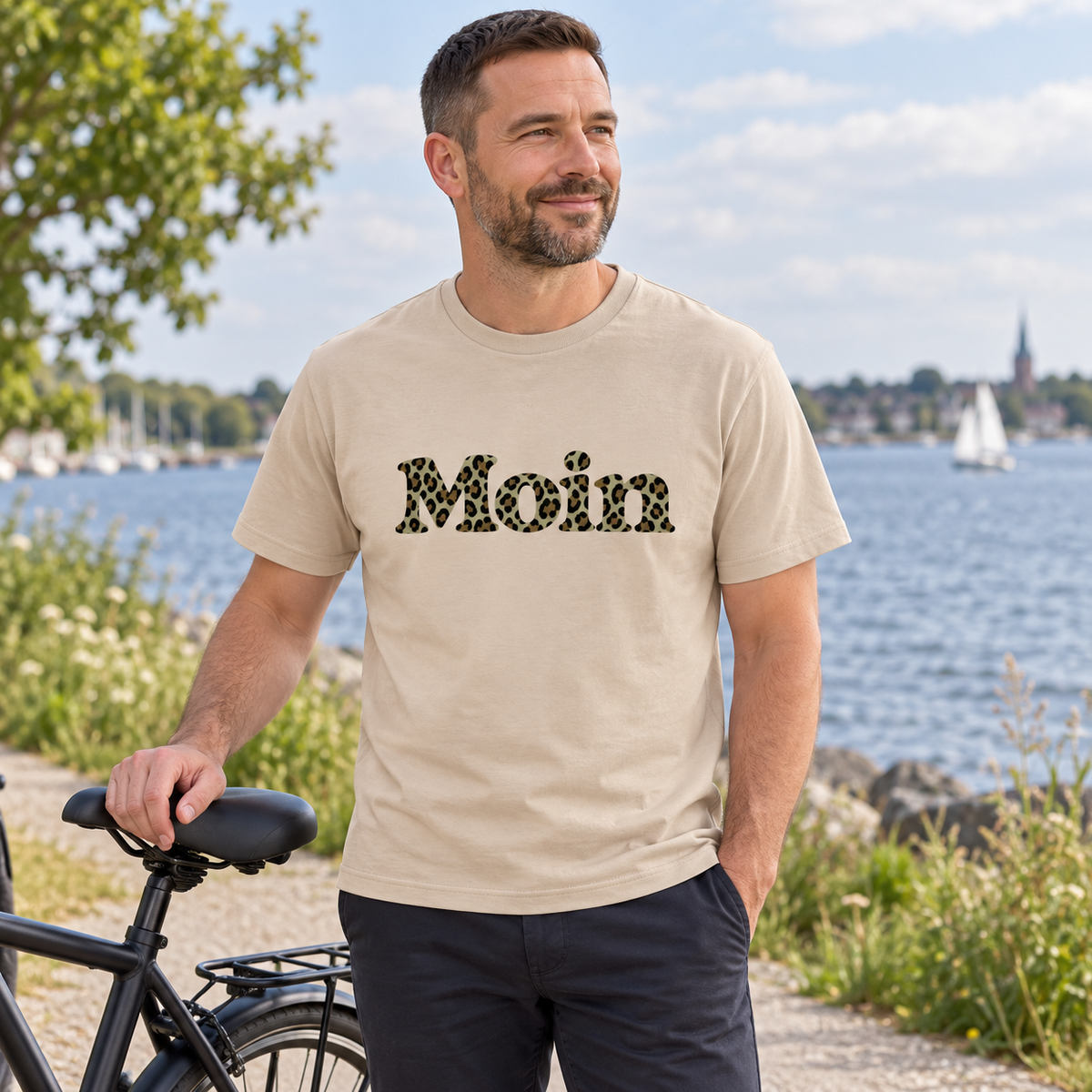 Mann im Essential Unisex T-Shirt in Sand mit Moin-Leoprint der Sage-Green-Edition als Frontprint neben einem Fahrrad