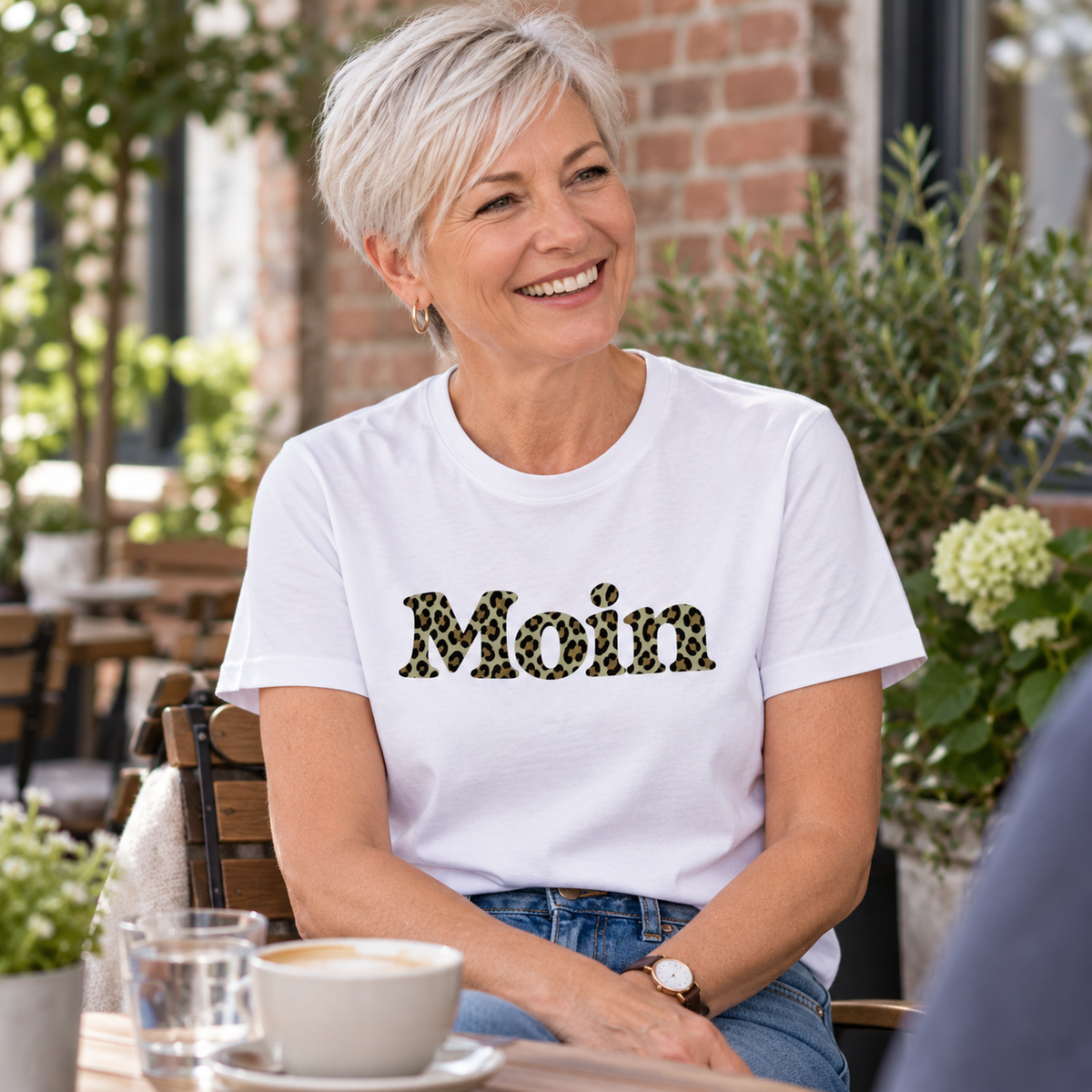 Frau im weißen Essential Unisex T-Shirt mit Moin-Leoprint der Sage-Green-Edition als Frontprint im Café