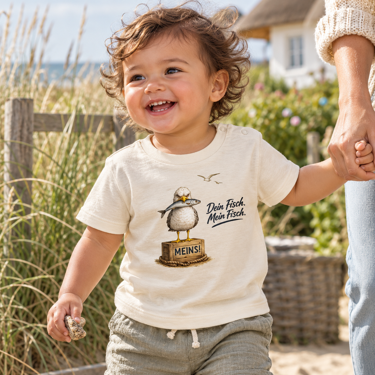 Kleinkind im Baby Bio T-Shirt Natural Raw mit Möwe, Fisch und Schriftzug als Frontprint im Küstengarten