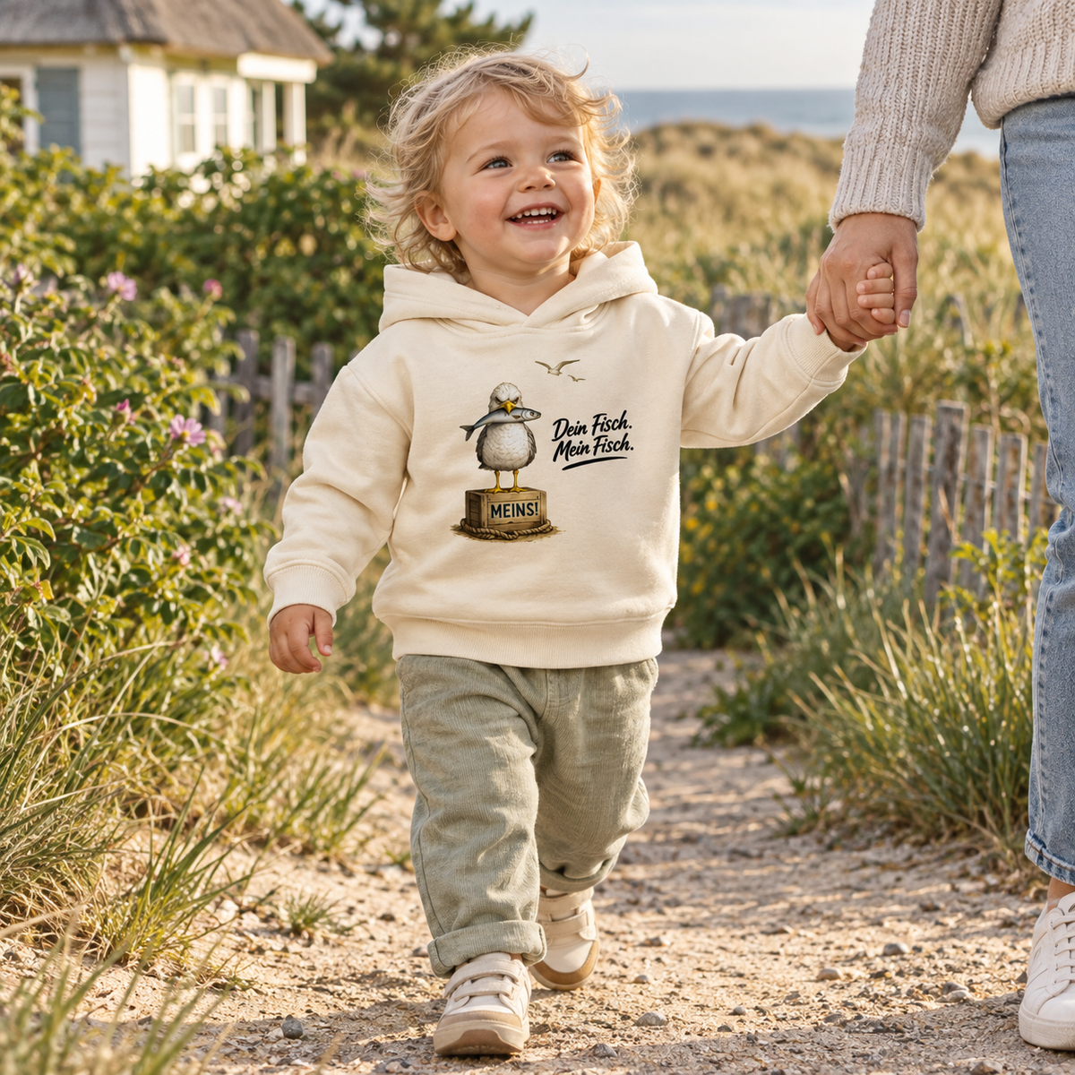 Kleinkind im naturfarbenen Baby Bio-Hoodie mit Fisch-Möwe als Frontprint im Küstengarten