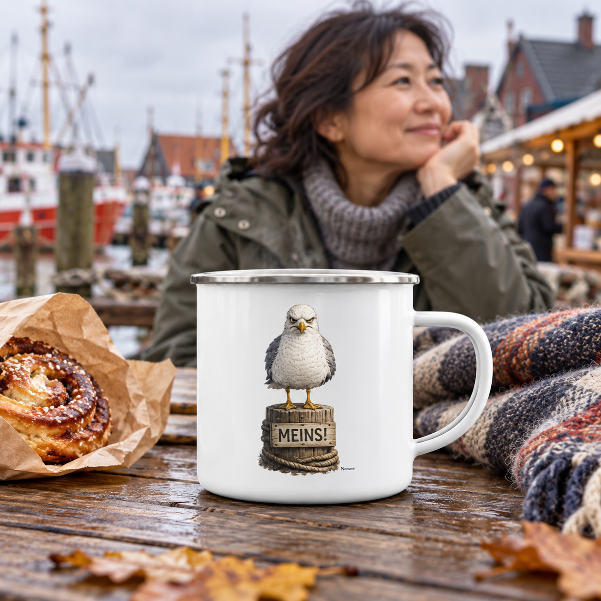 Weiße 300-ml-Emailletasse mit Silberrand und Alpha-Möwe auf „MEINS!“-Poller an einem Küstenhafenmarkt