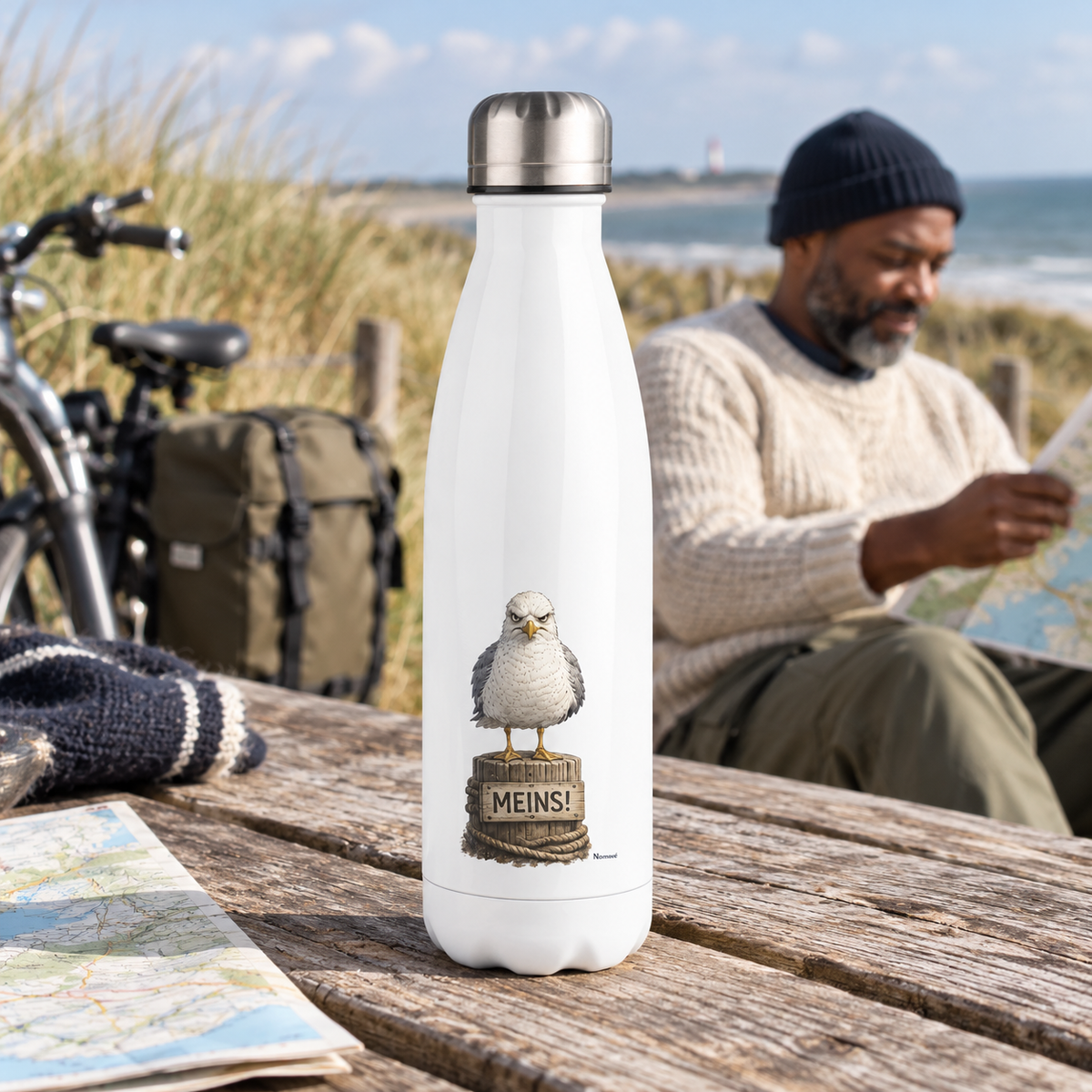 Weiße Edelstahl-Thermoflasche mit Möwe und „MEINS!“ auf einem Picknicktisch an der Küste