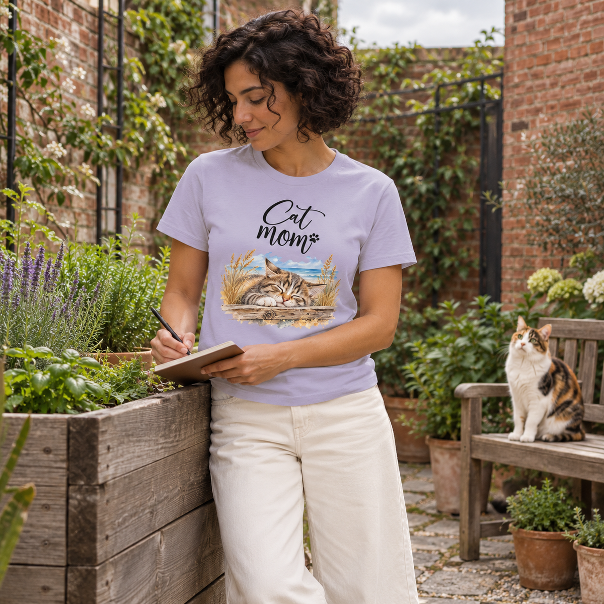 Frau im Damen-Bio-T-Shirt in Lavender mit Cat-Mom-Katzenmotiv vorne schreibt am Hochbeet; eine dreifarbige Katze sitzt auf der Gartenbank.