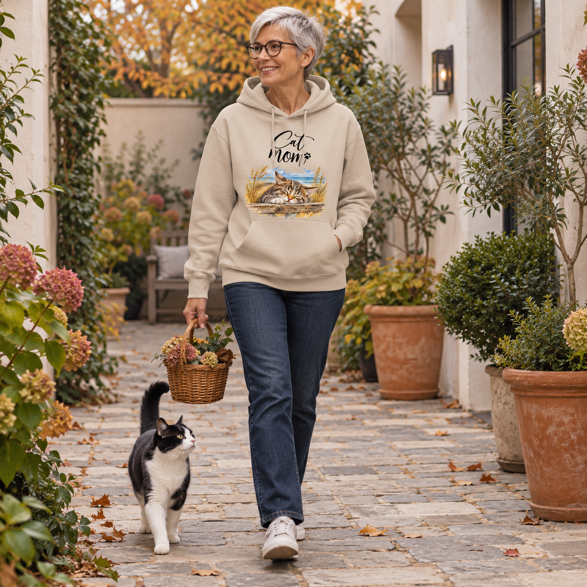 Frau im Bio-Hoodie in Desert Dust mit Cat-Mom-Katzenmotiv vorne geht mit einem Korb durch den Innenhof; eine schwarz-weiße Katze läuft neben ihr.