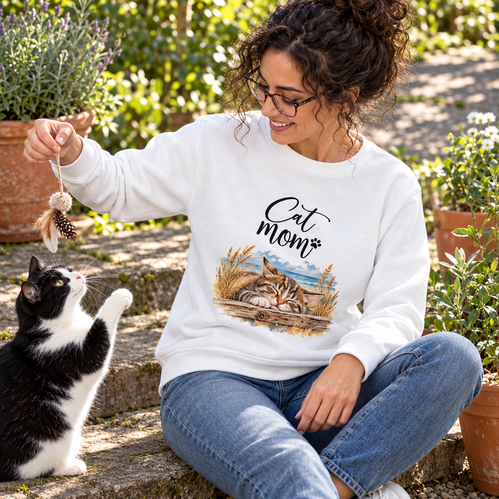 Frau im weißen Cat-Mom-Bio-Sweatshirt spielt im Garten mit einer schwarz-weißen Katze; Küstenmotiv als Frontprint.