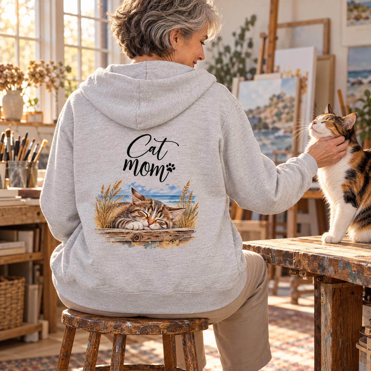 Frau im Bio-Zipper Heather Grey mit Cat-Mom-Backprint streichelt eine dreifarbige Katze im Atelier.