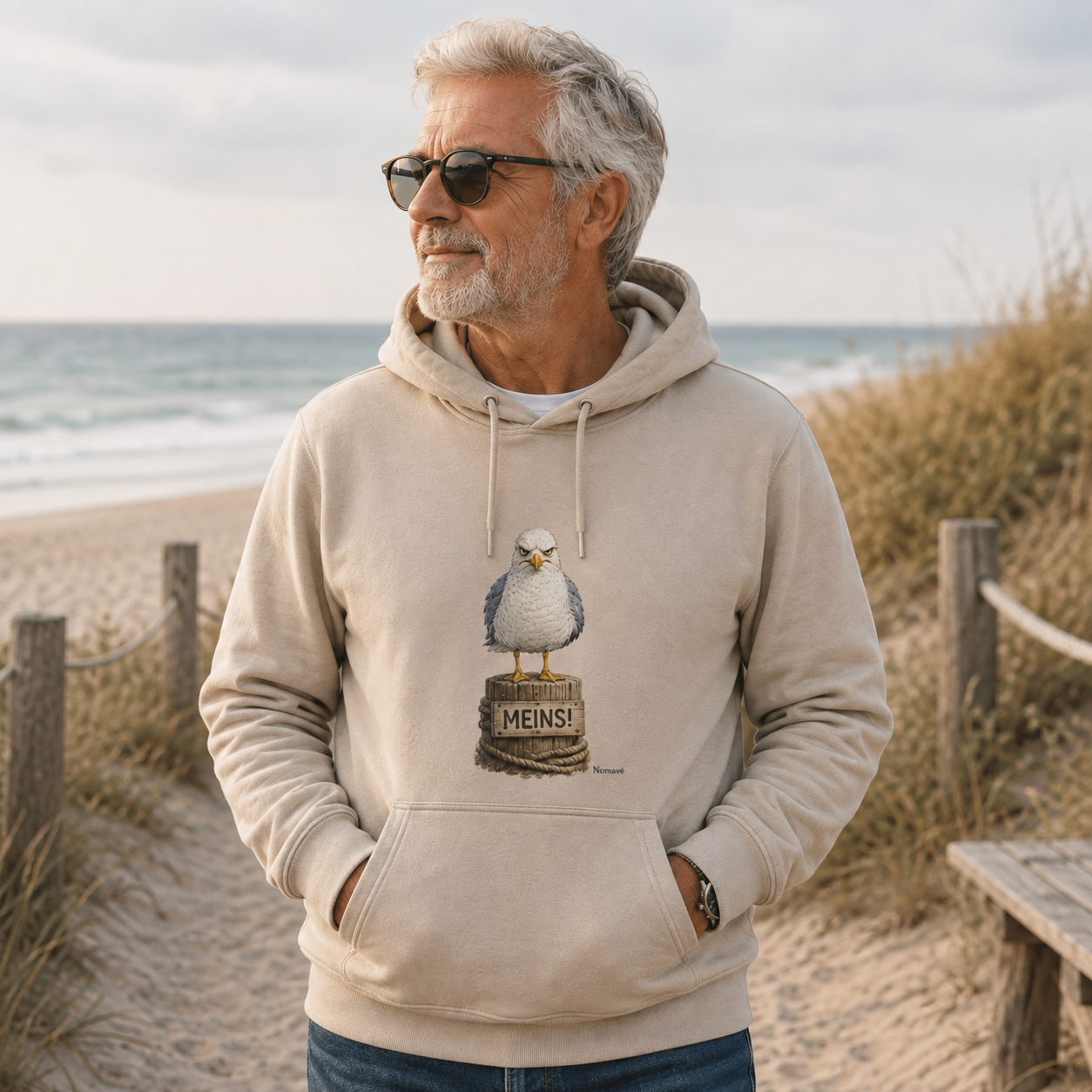 Älterer Mann trägt einen hellen Nomavé Hoodie mit dem Küstenhumor-Motiv „MEINS!“ und der frechen Möwe Alpha am Ostseestrand zwischen Dünen und Meer.