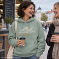 Kühlungsborn Baltic Waves – Bio-Hoodie, Dark Edition, Aloe – beim Warten am Kaffeekiosk mit einer Freundin, Frontdruck; KI-generierte Produktvisualisierung.