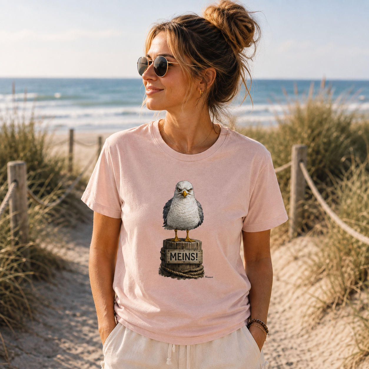 Junge Frau trägt das rosafarbene Bio T-Shirt „MEINS!“ aus der Nomavé Küstenhumor-Kollektion und steht entspannt auf einem Dünenweg am Ostseestrand mit Blick auf das Meer.